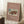 May include: Framed vintage Japanese woodblock print poster leaning against white wall beside cozy beige armchair and wooden side table with coffee cup. The artwork features a snow-covered traditional red temple gate with people and cart in winter landscape, displayed on dusty rose background with cream text. Natural lighting creates soft shadows on white fluffy rug.