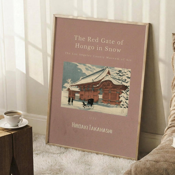 May include: Framed vintage Japanese woodblock print poster leaning against white wall beside cozy beige armchair and wooden side table with coffee cup. The artwork features a snow-covered traditional red temple gate with people and cart in winter landscape, displayed on dusty rose background with cream text. Natural lighting creates soft shadows on white fluffy rug.