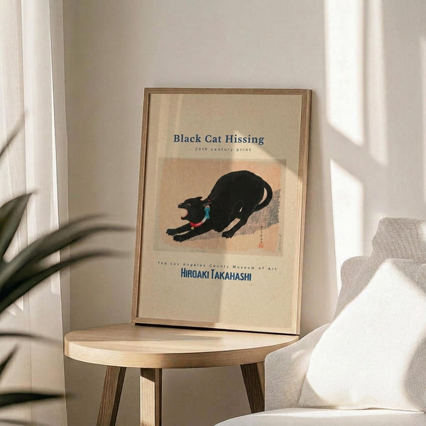 May include: Framed art print featuring a black cat in an arched, hissing pose with blue collar on cream and peach background. Museum poster format with text above and below the image. Displayed on round wooden side table next to beige upholstered chair with white pillow. Soft natural lighting creates shadows on white wall, with green plant leaves visible at left edge.