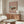 May include: Vintage Japanese woodblock print poster displayed above wooden sideboard in minimalist living room. Artwork shows traditional temple building with red gate in snowy winter scene, set against dusty rose background with white text. Modern furniture includes cream curved chair, glass oval coffee table, and wooden floor lamp. Natural lighting creates warm, contemporary atmosphere.