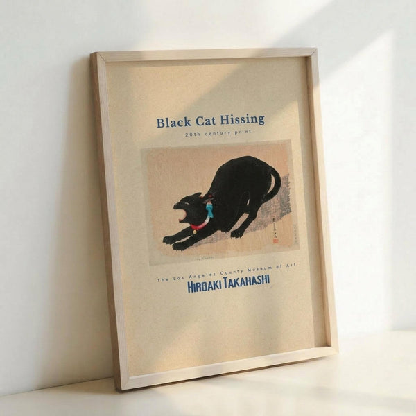 May include: Framed vintage Japanese woodblock print of a black cat in an arched, hissing pose with open mouth showing blue interior and red collar. The artwork features muted peach and brown tones with Japanese text. Light wood frame leans against white wall in bright, minimalist setting with visible shadow cast across the print.