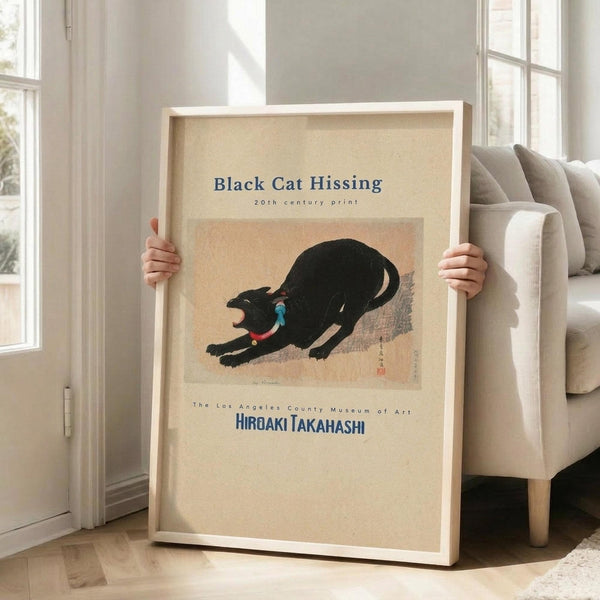 May include: Person holding a large framed poster in a bright living room with beige sofa and white walls. The vintage-style print features a black cat in a hissing pose with arched back on cream background, displayed in light wood frame. Natural sunlight streams through tall windows onto hardwood floors.