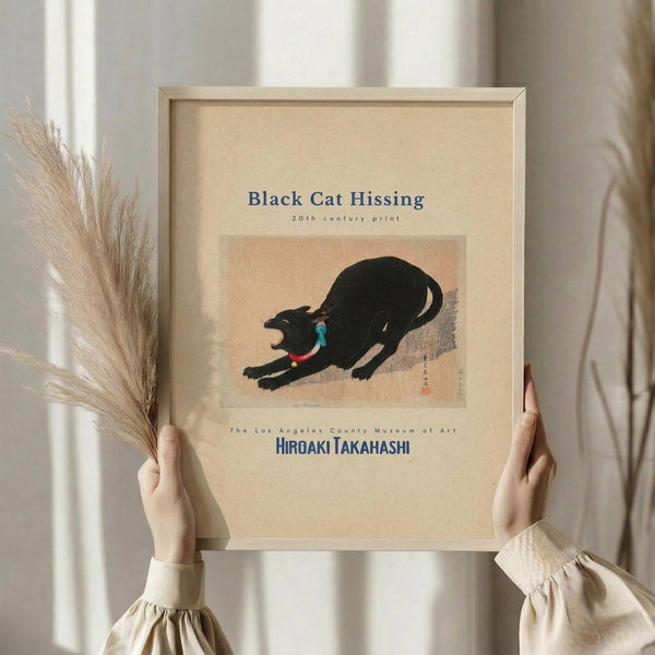 May include: Hands holding a framed art print featuring a black cat in an arched, hissing pose against a peach and cream background. The vintage-style Japanese print shows museum text and artist attribution. Natural wood frame with cream matting. Person wears beige clothing with gathered sleeves. Dried pampas grass visible to the left. Soft natural lighting against neutral curtained background.