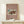 May include: Framed vintage Japanese woodblock print poster displayed on light wood console table against cream wall. Art shows traditional red temple building in snow with small figures and rickshaw, set on mauve background with white text. Natural wood frame, small wrapped gift with greenery sits beside poster.