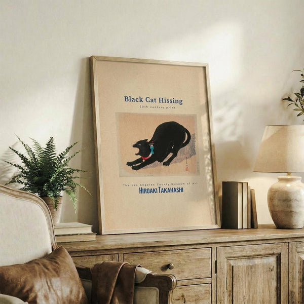 May include: Framed poster featuring a black cat in hissing pose with arched back and open mouth, displayed on rustic wooden sideboard alongside fern plant, stacked books, and ceramic pottery. The artwork shows Japanese-style illustration with blue and red collar details against beige background. Natural lighting creates warm ambiance in neutral-toned living room setting.