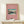 May include: Framed poster with pink background featuring Japanese woodblock print of snow-capped Mt. Fuji reflected in blue water, surrounded by trees under turquoise sky. Gold thin frame displayed on light wood surface alongside small white gift box tied with green sprig against cream wall.