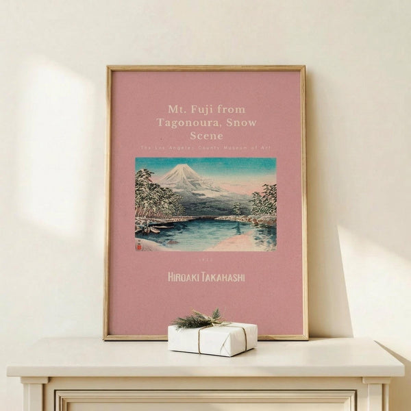 May include: Framed poster with pink background featuring Japanese woodblock print of snow-capped Mt. Fuji reflected in blue water, surrounded by trees under turquoise sky. Gold thin frame displayed on light wood surface alongside small white gift box tied with green sprig against cream wall.