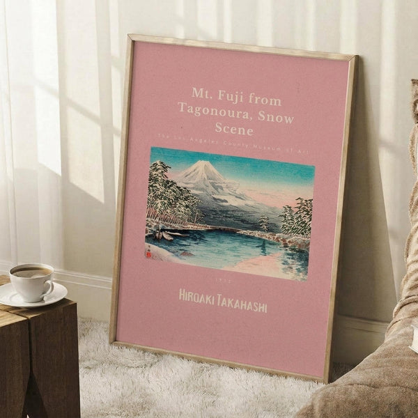 May include: Framed Japanese woodblock print poster featuring Mt. Fuji with snow-capped peak rising above a winter landscape with bare trees and a bridge over blue water, displayed against white paneled wall on white shag rug next to wooden side table with white coffee cup and cream-colored blanket