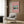 May include: Pink framed vintage Japanese woodblock print of Mount Fuji with snow-capped peak reflected in blue water, surrounded by trees under turquoise sky. Displayed on warm wood paneled wall above cream upholstered armchair with geometric patterned pillow. Modern brass floor lamp and dark round side table with open book complete the cozy reading nook scene.