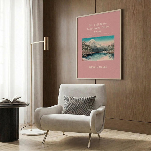 May include: Pink framed vintage Japanese woodblock print of Mount Fuji with snow-capped peak reflected in blue water, surrounded by trees under turquoise sky. Displayed on warm wood paneled wall above cream upholstered armchair with geometric patterned pillow. Modern brass floor lamp and dark round side table with open book complete the cozy reading nook scene.
