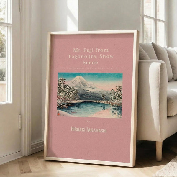 May include: Framed art print with coral pink background displaying a Japanese woodblock landscape of snow-capped Mt. Fuji viewed across a lake with evergreen trees. Print features museum exhibition styling with white text. Light wood frame leans against white wall beside a beige upholstered armchair in a bright room with tall windows and hardwood floors.