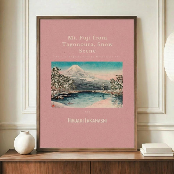 May include: Framed poster with pink background featuring vintage Japanese woodblock print of snow-capped Mt. Fuji, lake, and trees. White text shows artwork title and artist name Hiroaki Takahashi. Brown wooden frame sits on wooden credenza beside white ceramic vase and stacked books against cream walls with white molding.