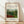 May include: Framed art print leaning against white wall on light wood floor. Print shows Henri Rousseau's lush jungle scene with dense green foliage, red flowers, and monkeys among tropical plants. Cream background with black text identifying the artwork and National Gallery of Art. Natural wood frame with warm lighting casting diagonal shadows across the wall.