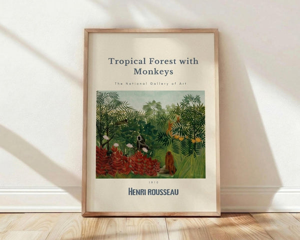 May include: Framed art print leaning against white wall on light wood floor. Print shows Henri Rousseau's lush jungle scene with dense green foliage, red flowers, and monkeys among tropical plants. Cream background with black text identifying the artwork and National Gallery of Art. Natural wood frame with warm lighting casting diagonal shadows across the wall.