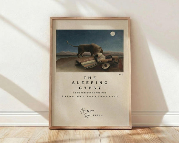 May include: Framed art print of Henri Rousseau's famous painting showing a sleeping figure in colorful striped clothing lying in a desert landscape with a lion standing nearby under a full moon. The poster features the title "The Sleeping Gypsy" and artist name below the artwork. Natural wood frame with cream matting, leaning against white wall on light wooden floor with soft window light casting shadows.