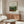 May include: Vintage art poster of tropical jungle scene with lush green foliage and red flowers, displayed in thin brown frame above wooden sideboard. Modern living room setting with cream upholstered chair, glass-top oval coffee table, and white table lamp. Neutral beige walls with natural lighting from window.
