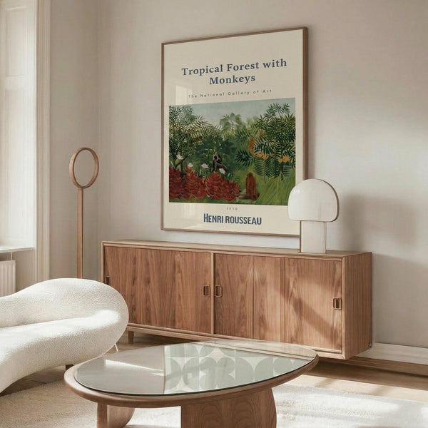 May include: Vintage art poster of tropical jungle scene with lush green foliage and red flowers, displayed in thin brown frame above wooden sideboard. Modern living room setting with cream upholstered chair, glass-top oval coffee table, and white table lamp. Neutral beige walls with natural lighting from window.