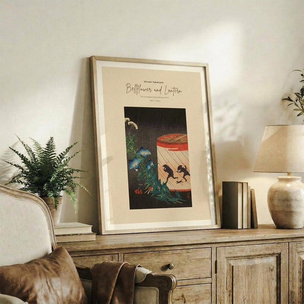 May include: Framed botanical art print displayed on rustic wooden console table in neutral living room setting. Print features dark floral design with blue morning glories, green foliage, and red paper lantern with black silhouettes against cream matting in light wood frame. Styled with fern plant, books, ceramic vase, and beige upholstered chair visible in cozy home decor arrangement.