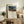 May include: Framed art print displaying a lion standing over a sleeping figure under a full moon in a desert landscape, with "THE SLEEPING GYPSY" text below the image and "Henry Rousseau" signature. Print sits on rustic wooden sideboard alongside fern plant, books, ceramic vase, and pottery jar. Cream upholstered chair with brown throw blanket visible in foreground against white wall.
