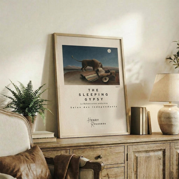 May include: Framed art print displaying a lion standing over a sleeping figure under a full moon in a desert landscape, with "THE SLEEPING GYPSY" text below the image and "Henry Rousseau" signature. Print sits on rustic wooden sideboard alongside fern plant, books, ceramic vase, and pottery jar. Cream upholstered chair with brown throw blanket visible in foreground against white wall.