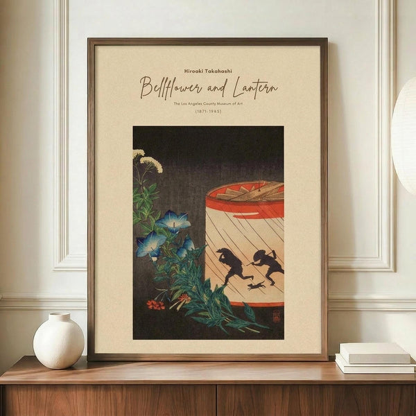 May include: Framed vintage Japanese woodblock print reproduction displayed on wooden sideboard against cream walls with white trim. Print shows blue morning glory flowers on left, red paper lantern with two black silhouetted figures running beneath on right, set against dark background. Cream colored mat, thin brown frame. Small white ceramic vase and stacked books visible on sideboard beside the artwork.