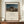 May include: Framed poster print displaying Henri Rousseau's 1897 painting featuring a sleeping figure in colorful striped clothing beside a lion under a full moon in a desert landscape. The cream matted print shows exhibition-style text below the artwork. Displayed on a wooden credenza with white ceramic vase and books in an elegant room with beige walls and white molding.