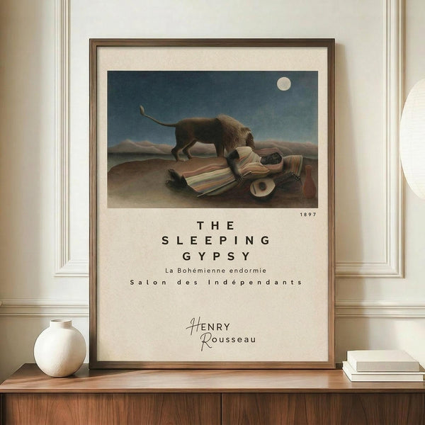 May include: Framed poster print displaying Henri Rousseau's 1897 painting featuring a sleeping figure in colorful striped clothing beside a lion under a full moon in a desert landscape. The cream matted print shows exhibition-style text below the artwork. Displayed on a wooden credenza with white ceramic vase and books in an elegant room with beige walls and white molding.
