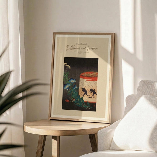 May include: Framed art print displayed on round wooden side table next to beige upholstered chair. Print shows Japanese-style illustration with bellflowers, red paper lantern, and silhouettes against dark background. Natural wood frame with cream matting. Soft natural lighting creates shadows on white wall. Green plant leaves visible in corner.