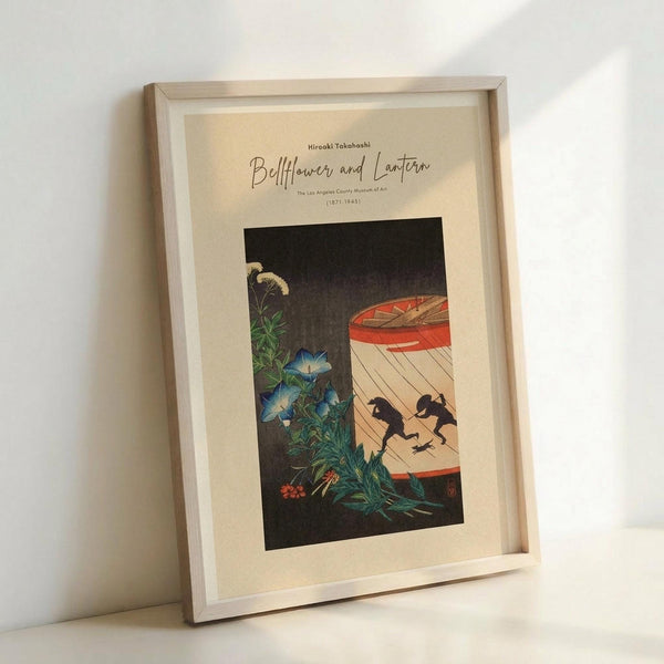 May include: Framed museum poster featuring Japanese woodblock print style artwork with blue bellflowers and green foliage on left side, red paper lantern with black silhouettes on right side, displayed in light wooden frame against white wall in bright interior setting