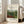 May include: Framed art print leaning against white wall next to beige sofa in bright living room. Print shows lush tropical jungle scene with green foliage, red flowers, and small monkeys among dense vegetation. Museum-style poster format with cream background, black text reading "Tropical Forest with Monkeys" and "Henri Rousseau" with "The National Gallery of Art" and "1910" details. Natural wood frame, hardwood floors visible.