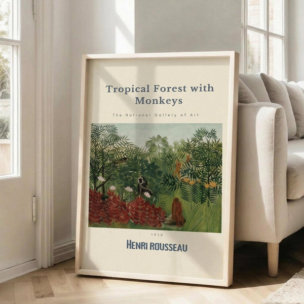 May include: Framed art print leaning against white wall next to beige sofa in bright living room. Print shows lush tropical jungle scene with green foliage, red flowers, and small monkeys among dense vegetation. Museum-style poster format with cream background, black text reading "Tropical Forest with Monkeys" and "Henri Rousseau" with "The National Gallery of Art" and "1910" details. Natural wood frame, hardwood floors visible.