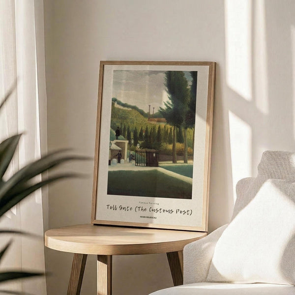 May include: A framed art print displayed on a round wooden side table next to a beige upholstered chair. The artwork shows a pastoral scene with green hills, trees, a small building with gates, and figures in muted earth tones. The print has a white mat border with text below the image and is housed in a thin wooden frame. Soft natural light creates shadows across the neutral-toned room setting, with a plant visible in the corner.