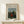 May include: Framed art print of Henri Rousseau's naive-style painting showing a woman in red dress seated in lush green landscape with church spire in background. Poster displayed on light wood surface with small white wrapped gift beside it. Thin gold frame against cream wall with soft natural lighting.
