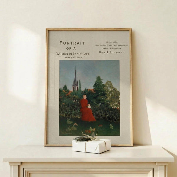 May include: Framed art print of Henri Rousseau's naive-style painting showing a woman in red dress seated in lush green landscape with church spire in background. Poster displayed on light wood surface with small white wrapped gift beside it. Thin gold frame against cream wall with soft natural lighting.