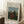 May include: Framed art print leaning against white wall corner showing woman in red dress sitting in lush green landscape with church spire in background. Light wood frame on white mat, positioned on textured cream rug next to wooden stool with white coffee cup and saucer.