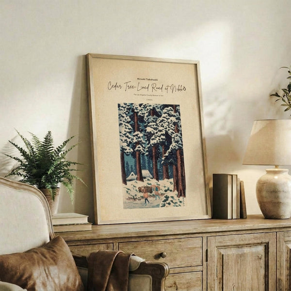 May include: Framed vintage Japanese woodblock print showing snow-covered cedar trees with small figures walking below, displayed on rustic wooden console table alongside fern plant, ceramic vase, and books in neutral-toned living room setting