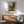 May include: Framed vintage-style landscape painting print hanging on white wall above wooden sideboard with table lamp. Green upholstered mid-century armchair with wooden arms in foreground. Artwork shows pastoral scene with gate, rolling hills, and trees in muted earth tones. Natural sunlight creates shadows across the minimalist interior setting.