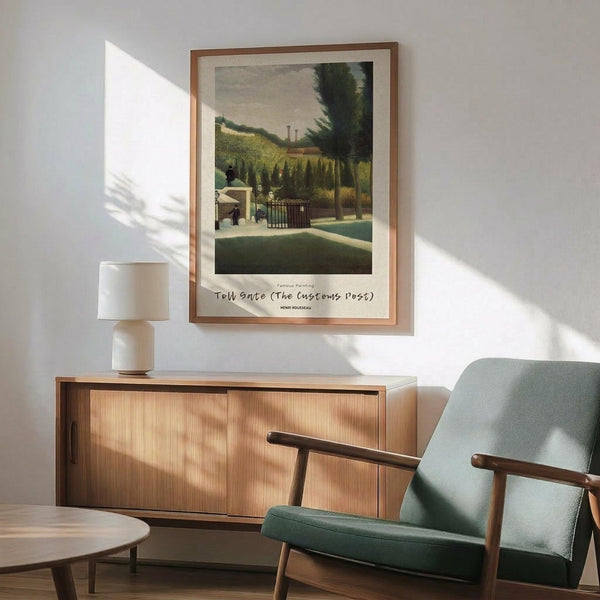 May include: Framed vintage-style landscape painting print hanging on white wall above wooden sideboard with table lamp. Green upholstered mid-century armchair with wooden arms in foreground. Artwork shows pastoral scene with gate, rolling hills, and trees in muted earth tones. Natural sunlight creates shadows across the minimalist interior setting.