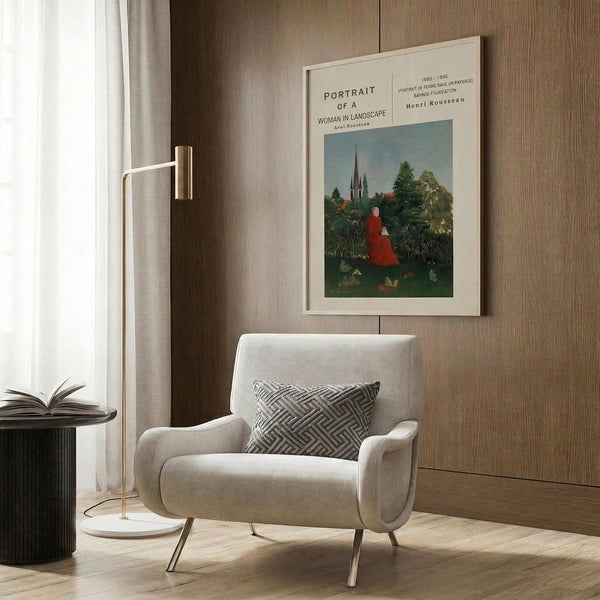 May include: Framed art print displayed on wood-paneled wall showing woman in red dress sitting in green landscape with church spire in background. Cream-colored frame mounted above beige upholstered armchair with geometric patterned pillow. Modern reading corner with brass floor lamp, side table with open book, and white curtains.