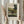 May include: Hands holding a framed art print of a vintage landscape painting showing a rural scene with green hills, trees, buildings with smokestacks, and figures near a gate. The print has a beige mat with handwritten-style text identifying it as a Henri Rousseau work. One hand also holds dried wheat or pampas grass stems. Neutral beige clothing and soft window lighting visible in background.