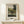 May include: Framed art print of a French Post-Impressionist painting showing a rural toll gate scene with a small house, iron gate, large trees, and rolling green hills in the background. The artwork features muted greens, yellows and earth tones in a naive folk art style. Light wooden frame with white matting, displayed on a cream wooden console table alongside a small white wrapped gift with twine and evergreen sprig against a neutral wall.