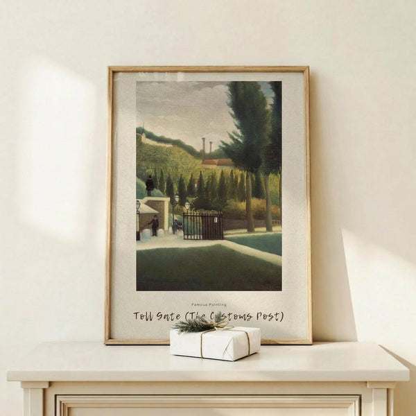 May include: Framed art print of a French Post-Impressionist painting showing a rural toll gate scene with a small house, iron gate, large trees, and rolling green hills in the background. The artwork features muted greens, yellows and earth tones in a naive folk art style. Light wooden frame with white matting, displayed on a cream wooden console table alongside a small white wrapped gift with twine and evergreen sprig against a neutral wall.