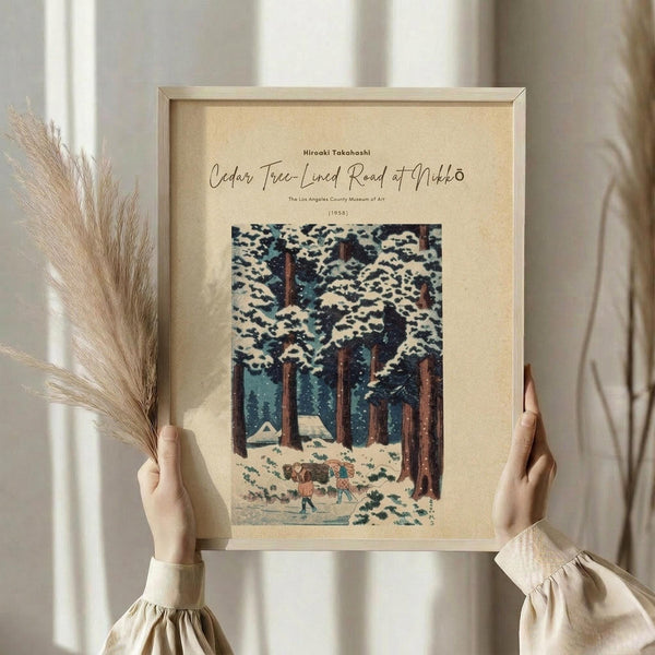 May include: Hands holding a framed vintage Japanese woodblock print showing snow-covered cedar trees with small figures walking below, displayed in light wood frame with cream matting. Dried wheat stems visible to the left. Soft natural lighting creates gentle shadows on neutral beige wall background.