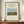 May include: Framed poster print featuring Henri Rousseau's pastoral painting with a brown and white cow in a green meadow, haystacks, trees, and church spire under blue sky. Black text reads "Scene in Bagneux on the Outskirts of Paris 1909" with artist signature below. Thin brown wooden frame displayed on walnut credenza beside white ceramic vase and stacked books against cream wainscoted wall.