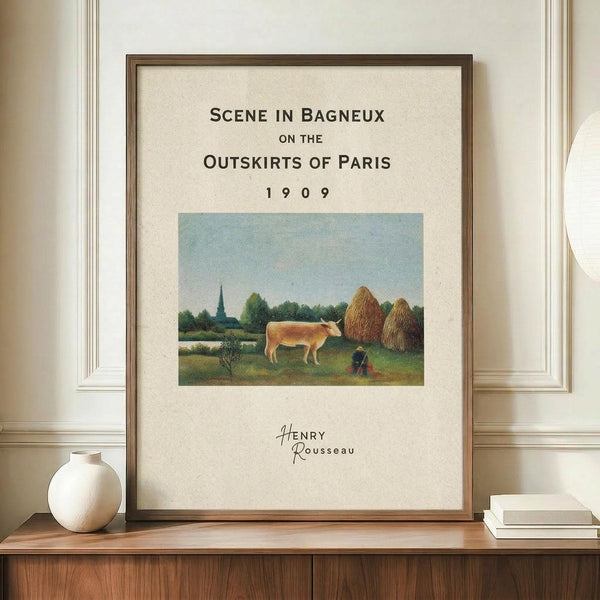 May include: Framed poster print featuring Henri Rousseau's pastoral painting with a brown and white cow in a green meadow, haystacks, trees, and church spire under blue sky. Black text reads "Scene in Bagneux on the Outskirts of Paris 1909" with artist signature below. Thin brown wooden frame displayed on walnut credenza beside white ceramic vase and stacked books against cream wainscoted wall.