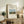 May include: Framed art print displaying a pastoral landscape with a brown cow, haystacks, and church spire under blue sky, positioned on rustic wooden sideboard alongside fern plant, ceramic vase, and books in warm-toned living room setting with beige upholstered chair visible.