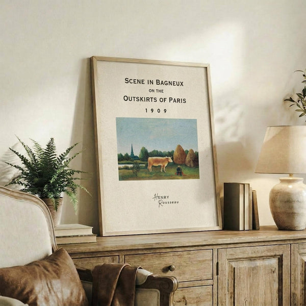 May include: Framed art print displaying a pastoral landscape with a brown cow, haystacks, and church spire under blue sky, positioned on rustic wooden sideboard alongside fern plant, ceramic vase, and books in warm-toned living room setting with beige upholstered chair visible.