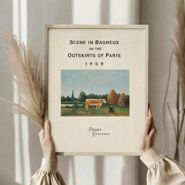 May include: Hands holding a framed art print featuring a pastoral scene with a brown and white cow, haystacks, and green countryside. The print has cream matting with black text reading "Scene in Bagneux on the Outskirts of Paris 1909" and "Henry Rousseau" below the artwork. Person wearing a cream-colored ruffled sleeve top holds dried pampas grass in one hand while displaying the wooden-framed poster against a soft beige background with natural window lighting.