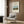 May include: Framed vintage art poster featuring Henri Rousseau's pastoral painting with cow in countryside landscape, displayed on wood-paneled wall above beige upholstered chair with geometric patterned pillow, brass floor lamp and dark round side table with open book nearby