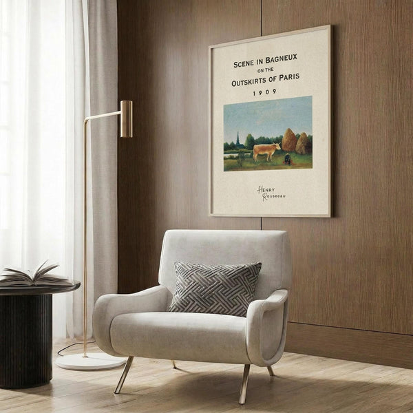May include: Framed vintage art poster featuring Henri Rousseau's pastoral painting with cow in countryside landscape, displayed on wood-paneled wall above beige upholstered chair with geometric patterned pillow, brass floor lamp and dark round side table with open book nearby